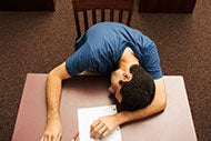 A person sleeps at a table. There is a piece of paper and pen on the desk