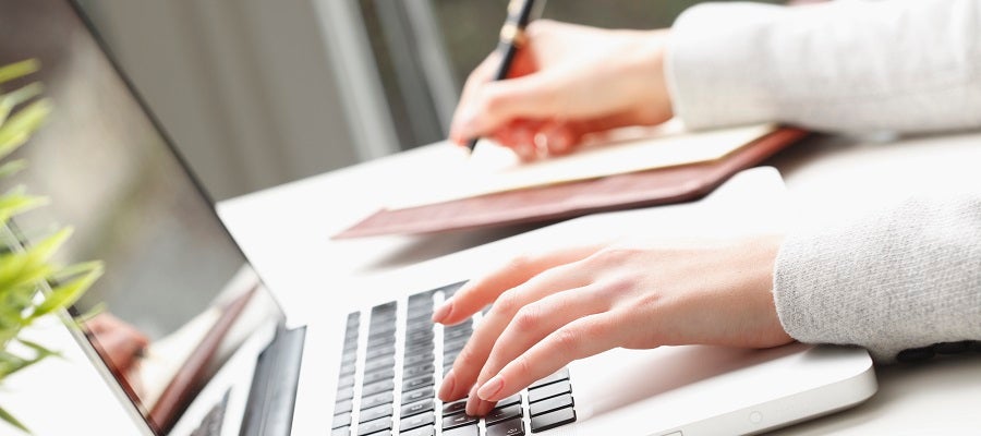 A woman types on a laptop computer with her left hand as the takes notes in a notebook using a pen with her right hand.